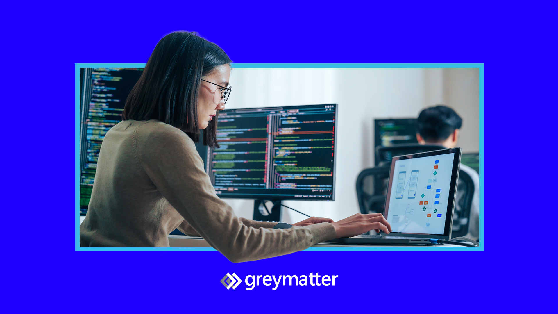 Developer in office working on laptop, with monitors in the background displaying code. Grey Matter logo at the bottom